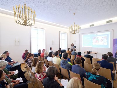 European Year of Skills wie Europass die systematische Dokum Das Bild zeigt eine Europass Veranstaltung. Eine weibliche Person präsentiert vor einem Pult, Veranstaltungsteilnehmer und Teilnehmerinnen hören zu.