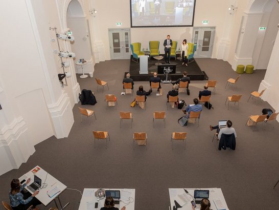 Bologna-Tag 2020. Think Big: Institutionelle Lehrstrategien und Ein großer Saal von schräg oben, im Hintergrund eine Bühne mit zwei Personen, davor Personen auf Stühlen mit Abstand
